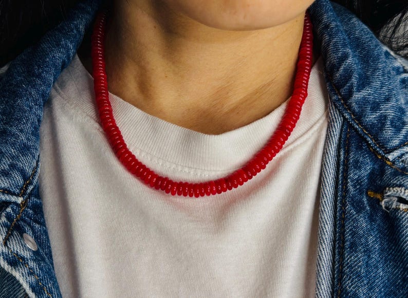 May include: A vibrant red necklace with a spiral design. The necklace is worn over a white t-shirt and a denim jacket. The necklace is a bold accessory.