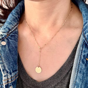 Könnte beinhalten: Eine goldfarbene Y-förmige Halskette mit einer filigranen Kette und einem kreisförmigen Anhänger. Die Halskette wird über einem grauen Oberteil und einer Jeansjacke getragen.