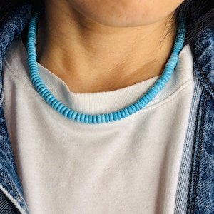 May include: A light blue beaded necklace resting on a white t-shirt and denim jacket. The necklace is composed of small, round beads, creating a continuous strand. The overall aesthetic is casual and stylish.