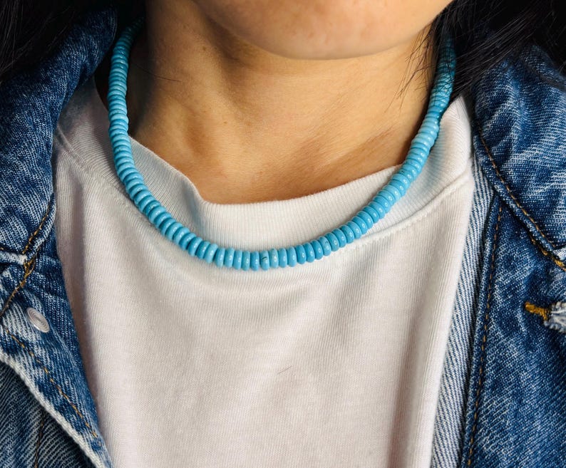 May include: A light blue beaded necklace resting on a white t-shirt and denim jacket. The necklace is composed of small, round beads, creating a continuous strand. The overall aesthetic is casual and stylish.