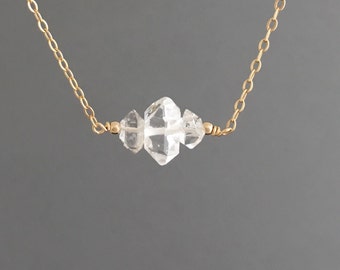 May include: A delicate gold chain necklace featuring three clear, faceted crystal stones. The crystals are clustered in the center of the necklace, with a small gold bead on each side. The necklace is displayed against a gray background.