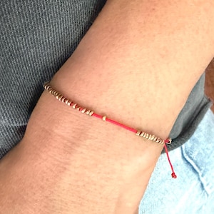 May include: A red string bracelet with small gold beads. The bracelet is tied with a knot and has a tassel.
