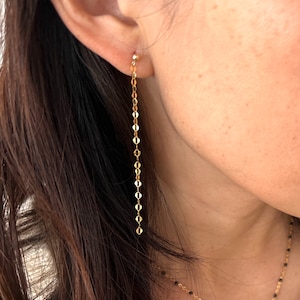 May include: Gold chain earrings with a small ball at the top. The earrings are long and dangle down to the wearer's neck.