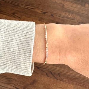 Può includere: Un delicato braccialetto d'oro con piccole perline alternate dorate e argentate. Il braccialetto è indossato su un polso, con una manica grigia visibile sullo sfondo. Il braccialetto è un gioiello semplice ed elegante.