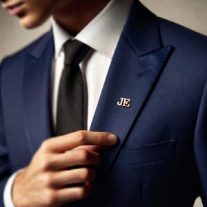 May include: A navy blue suit jacket with a white collared shirt and black tie. A gold-colored lapel pin with the initials "JE" is affixed to the jacket. The image focuses on the upper body and the pin.