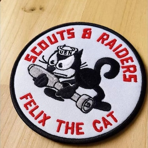 May include: A circular embroidered patch with a white background and a black border. It features a black and white cartoon cat wearing a sailor hat with the text U.S.N. The cat holds a gray object. Red text reads SCOUTS & RAIDERS FELIX THE CAT.