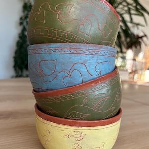 Peut inclure: Une pile de quatre bols en céramique avec un bord couleur terre cuite. Le bol supérieur est vert olive avec des motifs de cœurs, le deuxième est bleu avec des cœurs, le troisième est vert olive avec des feuilles, et le bas est jaune avec des animaux.