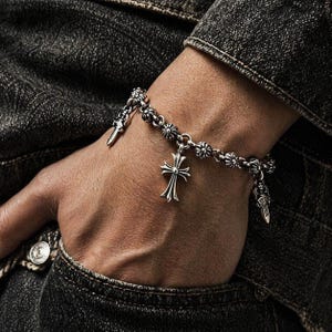 以下が含まれることがあります： クロス ペンダントと装飾的な短剣チャームが付いたシルバー ブレスレット。 花の形をしたリンクで作られたブレスレットは、濃い色のデニムジャケットを背景に手首に着用されています。