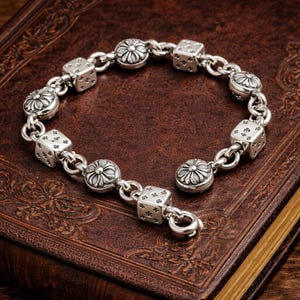 May include: Silver bracelet featuring alternating dice and flower-shaped charms. The dice have engraved dots, and the flower charms have a detailed petal design. The bracelet is connected by a chain and has a clasp closure. A stylish accessory.
