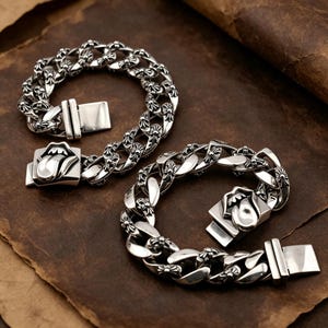 Può includere: Due braccialetti a catena argentati con un design a lingua e labbra sulla chiusura. I braccialetti hanno un design a catena a maglia grossa con accenti floreali decorativi. Presentati su una superficie marrone strutturata.