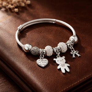 Op de afbeelding: Zilveren bedelarmband met meerdere decoratieve kralen en bedels. Bedels zijn onder meer een hart met de inscriptie "I love you", een teddybeer en een strik. De armband ligt op een bruin oppervlak.