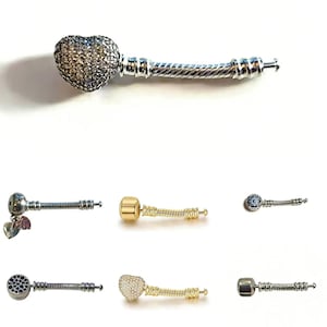 May include: A collection of charm bracelets with various designs. One charm features a heart shape covered in small, clear stones. Other charms include a gold-colored charm with a heart, a gold-colored charm with a round top, and charms with blue and black stones.