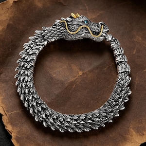 Op de afbeelding: Een zilveren drakenarmband met gedetailleerde schubben. De drakenkop heeft gouden accenten en een blauwe edelsteen in het oog. De armband is ontworpen om de pols te omsluiten, met een gedetailleerd, getextureerd ontwerp. Een uniek sieraad.