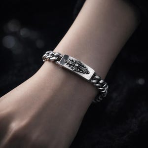 May include: Silver curb chain bracelet with a rectangular plaque bearing a floral design. The bracelet, with its chunky links, is shown on a wrist. The jewellery is set against a dark background. The bracelet's length is approximately 20 cm.