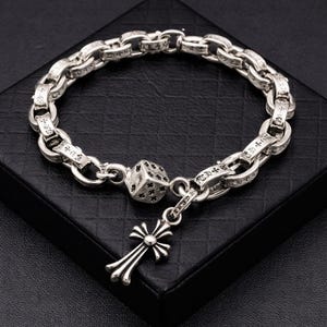 May include: Silver bracelet with a chain link design. The bracelet has a dice charm and a cross pendant. The links are engraved with text. The bracelet is displayed on a black surface.