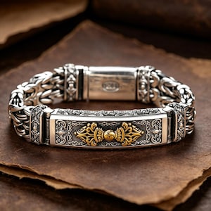 Puede incluir: Pulsera de plata con intrincados eslabones de cadena y una pieza central rectangular. La pieza central presenta diseños dorados ornamentados. La pulsera se muestra sobre una superficie marrón texturizada.