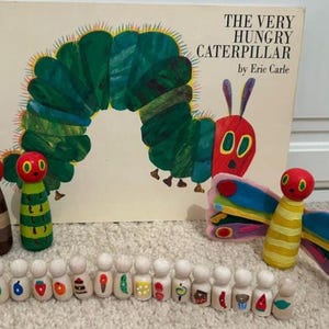 May include: A copy of "The Very Hungry Caterpillar" book by Eric Carle, with wooden peg dolls depicting the caterpillar and butterfly. Additional peg dolls are decorated with food items from the story. The book and dolls are arranged on a light-colored surface.
