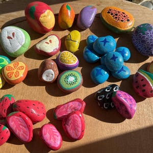 May include: A collection of smooth river stones hand-painted to resemble various fruits including strawberries, blueberries, kiwi, citrus slices, watermelon, and papaya. The stones feature vibrant colours and detailed patterns representing fruit textures.