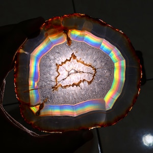 May include: A translucent, circular agate slice with a rainbow effect. The stone features concentric bands of color, including orange, yellow, green, and blue. The center has a crystalline structure with a brown outline. The edges are a reddish-brown color.