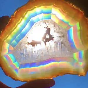 May include: A translucent agate slice featuring concentric bands of vibrant orange, yellow, and blue hues. The center contains a jagged, crystalline cavity that glows brightly when backlit, displaying a spectrum of iridescent rainbow colors.
