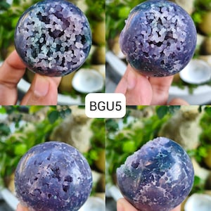 May include: A spherical purple agate stone featuring a central druzy geode cavity with small, crystalline formations. The surface displays deep violet and lavender hues with translucent, bubbly textures throughout. The text BGU5 is visible.