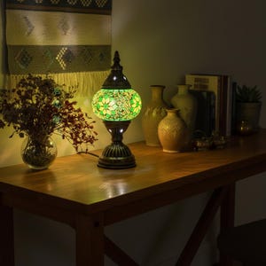 Peut inclure: Lampe de table décorative avec un abat-jour en mosaïque de verre vert orné de motifs floraux. La lampe, avec son socle en bronze foncé, est allumée et diffuse une lumière chaleureuse. Elle est posée sur une table en bois.