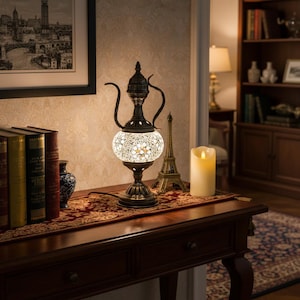 Könnte beinhalten: Eine dekorative Tischlampe mit einem Mosaikglasschirm und einem Sockel aus bronzefarbenem Metall. Die Lampe hat ein einzigartiges teekannenartiges Design mit gebogenen Griffen. Sie steht auf einem Holztisch mit rotem Muster.