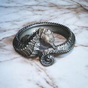 Può includere: Un bracciale rigido in metallo color argento con un design dettagliato a forma di cavalluccio marino. Il corpo del cavalluccio marino forma la fascia circolare, con la testa e la coda arricciata che si incontrano sull'apertura frontale.