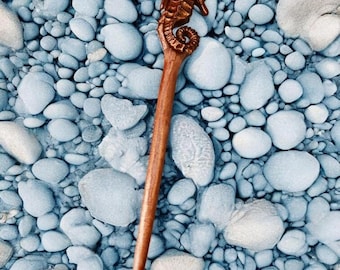 May include: A wooden hair stick with a seahorse carving at the top. The hair accessory is brown and has a long, slender design. The background is a collection of blue and white stones.