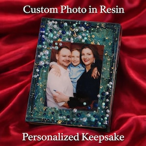 Pode incluir: Uma lembrança personalizada com uma foto de família embutida em resina. A moldura é decorada com contas azuis e brancas e flocos de ouro. O texto "Custom Photo in Resin" e "Personalized Keepsake" são exibidos.