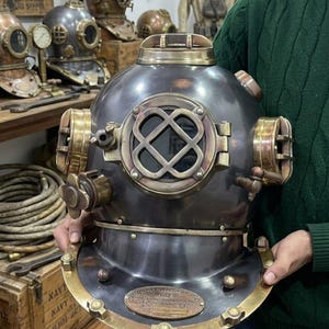 Könnte beinhalten: Ein Vintage-Tauchhelm mit dunkelgrauem Finish und Messingakzenten. Der Helm hat eine große, runde Frontplatte mit einem kreuzförmigen Fenster und mehreren Messingbeschlägen. Weitere Tauchhelme sind im Hintergrund zu sehen.