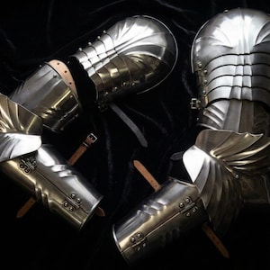 Puede incluir: Un conjunto de armadura de brazo de acero pulido, con placas articuladas y elementos decorativos en forma de ala. La armadura incluye correas y hebillas de cuero para sujetar, sobre un fondo oscuro.