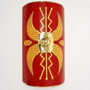 May include: A rectangular, curved Roman scutum shield featuring a vibrant red background. The design includes golden yellow wings, vertical zigzag arrows, and small rectangular accents. A polished brass central boss is mounted in the middle.