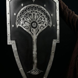 May include: A black shield with silver edging and a detailed tree design. The tree features a circular pattern and a long trunk. The shield is held by a person, with the design reminiscent of a fantasy theme.