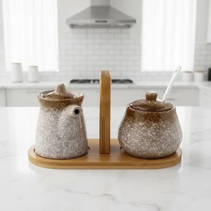 以下が含まれることがあります： 薄茶色と白の斑点模様のセラミックキッチンセット。セットには、小さなピッチャー、蓋とスプーン付きのジャー、ハンドル付きの木製トレイが含まれています。キッチン用品は調味料の提供を目的としています。