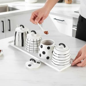 Puede incluir: Un conjunto de cuatro botes de cerámica blanca con diseños negros, incluyendo rayas, cuadros y lunares, cada uno con un asa negra en la tapa. Los botes están colocados sobre una bandeja rectangular blanca.