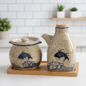 May include: A ceramic kitchen set featuring a speckled beige finish with blue fish and wave designs. The set includes a lidded jar and a spout bottle, both on a bamboo tray. The bottle has a wooden stopper.