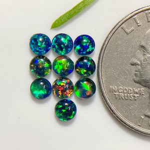 May include: Ten small, round, cabochon-cut synthetic opals arranged in a cluster. Each stone displays a dark base with vibrant flashes of blue, green, and orange fire. A United States quarter coin is placed next to the stones for size reference.