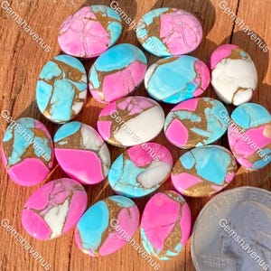 Puede incluir: Una colección de cabujones ovalados con un patrón jaspeado de segmentos de color rosa brillante, azul claro y blanco separados por vetas de color oro metálico. Se incluye una moneda para mostrar la escala y el tamaño de las piedras.