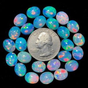 May include: A collection of twenty-two oval-shaped cabochon opals with a translucent blue base and vibrant flashes of green, orange, and pink, arranged around a silver-coloured United States quarter coin for scale on a solid black background.