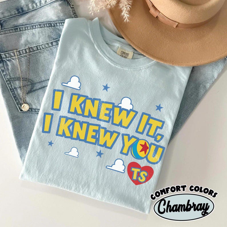 Pode incluir: Uma t-shirt azul-clara com o texto amarelo e azul I KNEW IT, I KNEW YOU, um cora&ccedil;&atilde;o vermelho com as letras TS, rodeado por pequenas nuvens brancas e estrelas azuis. A t-shirt &eacute; exibida com cal&ccedil;as de ganga azuis e um chap&eacute;u bege.