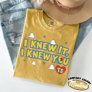 Pode incluir: Uma t-shirt amarelo-mostarda com o texto azul I KNEW IT, I KNEW YOU, um cora&ccedil;&atilde;o vermelho com as letras TS, gr&aacute;ficos de nuvens brancas e estrelas azuis. A t-shirt tem uma etiqueta Comfort Colors no colarinho e &eacute; de tamanho XL.