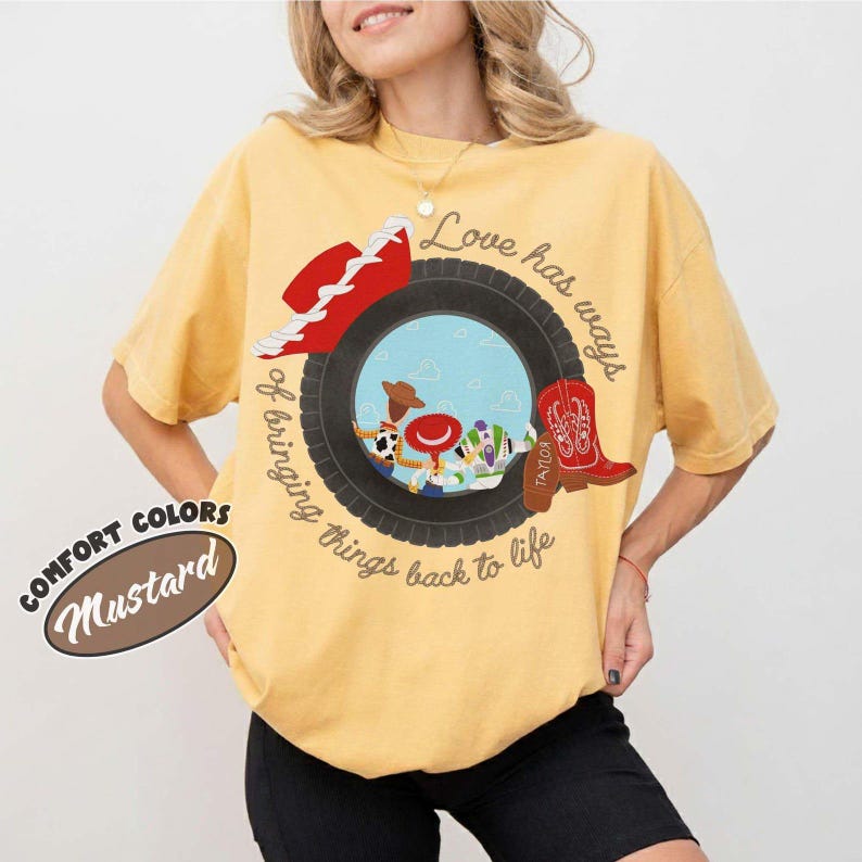 以下が含まれることがあります： マスタードイエローのTシャツで、黒いタイヤが青い空と漫画のキャラクターのシーンを囲むグラフィックが描かれています。デザインには赤いカウボーイハット、赤いカウボーイブーツ、そして「Love has ways of bringing things back to life」というテキストが含まれています。