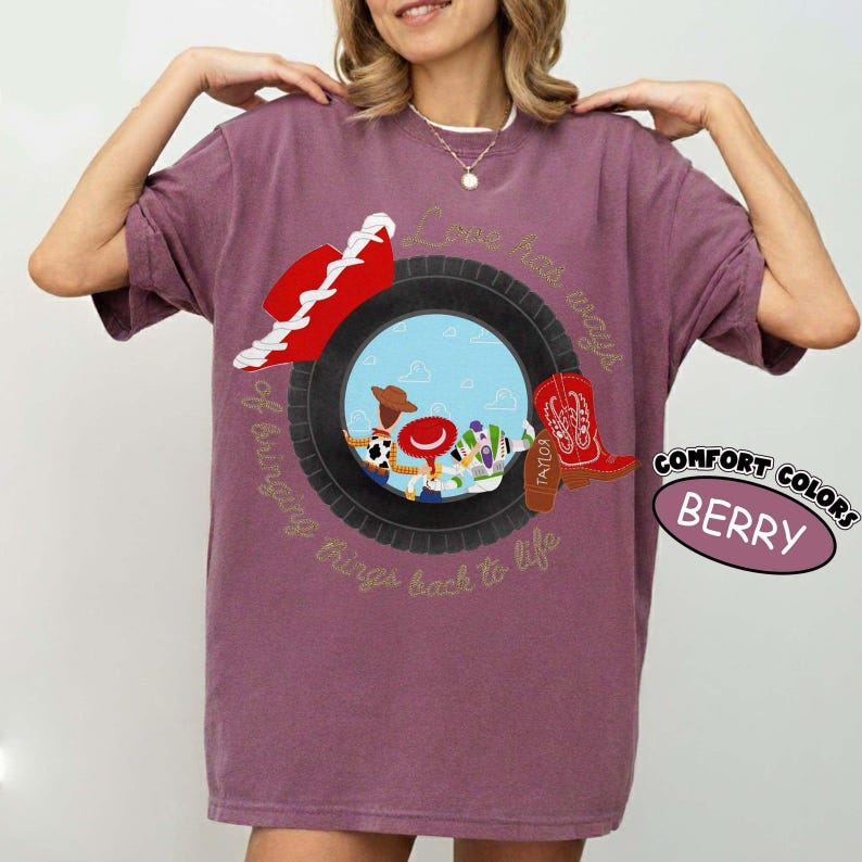 以下が含まれることがあります： ベリー色のTシャツで、黒いタイヤが3人の漫画キャラクターがいる青い空のシーンを囲むグラフィックが特徴です。赤いカウボーイハットと赤いカウボーイブーツがタイヤの上に配置されています。テキストには、Love has ways of bringing things back to lifeと書かれています。