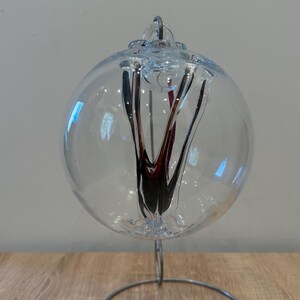 May include: A clear glass orb with dark red and black interior design, displayed on a silver metal stand. The orb has a decorative top and is set on a wooden surface. The design is abstract and artistic.