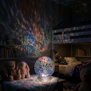 May include: A spherical glass lamp with a mosaic pattern of blue, red, and white shards. The lamp sits on a clear circular base and projects a vibrant, rippling light effect onto the surrounding wall, creating a colourful display of light.