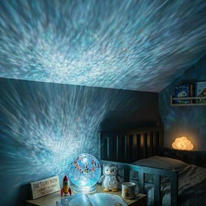 Può includere: Una lampada sferica con finitura in vetro crepato blu e bianco proietta un motivo luminoso ondulato sulla parete e sul soffitto. La lampada è appoggiata su un comodino in legno accanto a un libro aperto, un piccolo razzo giocattolo, un gufo di peluche e una tazza.