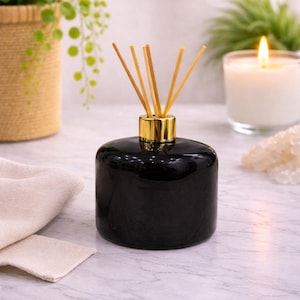May include: A black glass essential oil diffuser with a gold-colored collar, topped with wooden reeds. The diffuser sits on a marble surface, with a lit candle and greenery in the background.