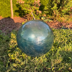 Op de afbeelding: Een bolvormige tuinkogel gemaakt van doorschijnend blauwgroen glas. Het oppervlak heeft een duidelijke gebarsten textuur over de hele bol, die het licht reflecteert met een glanzende afwerking. Het object heeft een gladde, ronde vorm.