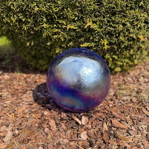May include: An iridescent gazing ball with a cracked glass effect. The sphere has a metallic finish with shades of blue, purple, and silver. It is resting on brown mulch in front of a green bush.